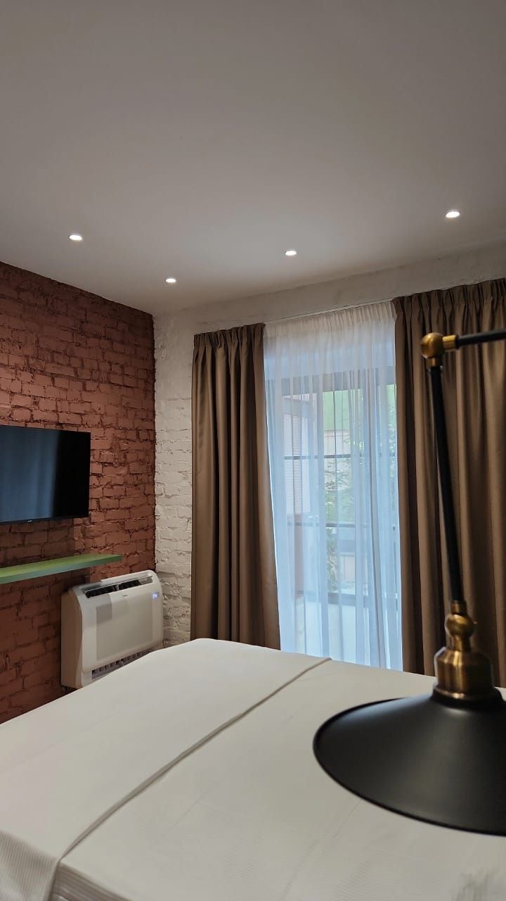 A hotel room with a brick wall, a flat-screen TV mounted on it, a bed with a white bedspread, a bedside table, a lamp, and a window with curtains.
