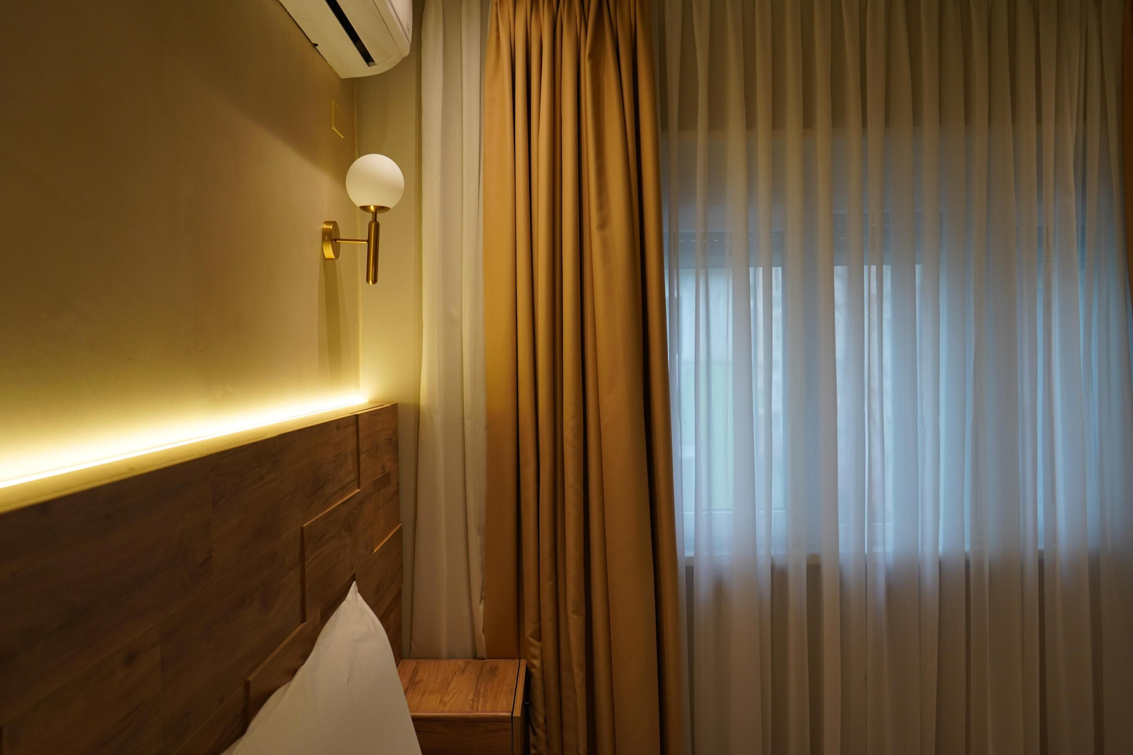 Ein Hotelzimmer mit hölzernem Kopfteil, einem Nachttisch, einer Lampe und einem Fenster mit transparenten Vorhängen.