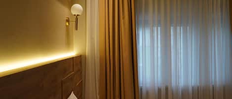 Une chambre d’hôtel avec une tête de lit en bois, une table de chevet, une lampe et une fenêtre avec des rideaux légers.