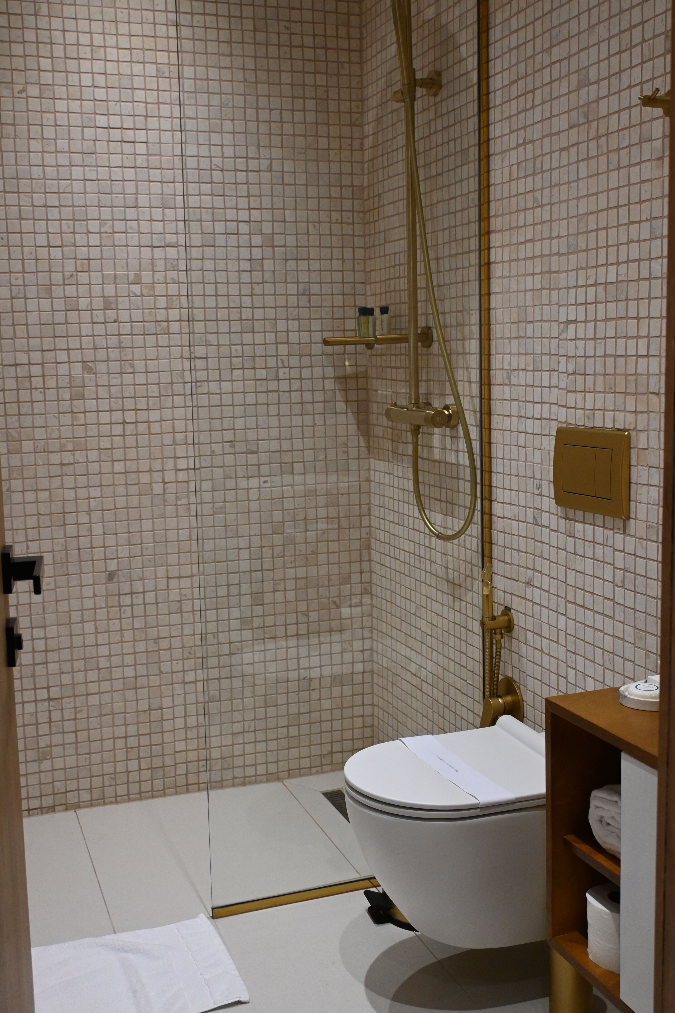 Un baño con pared de azulejos blancos, un inodoro blanco, una ducha con detalles dorados y un estante de madera con toallas.