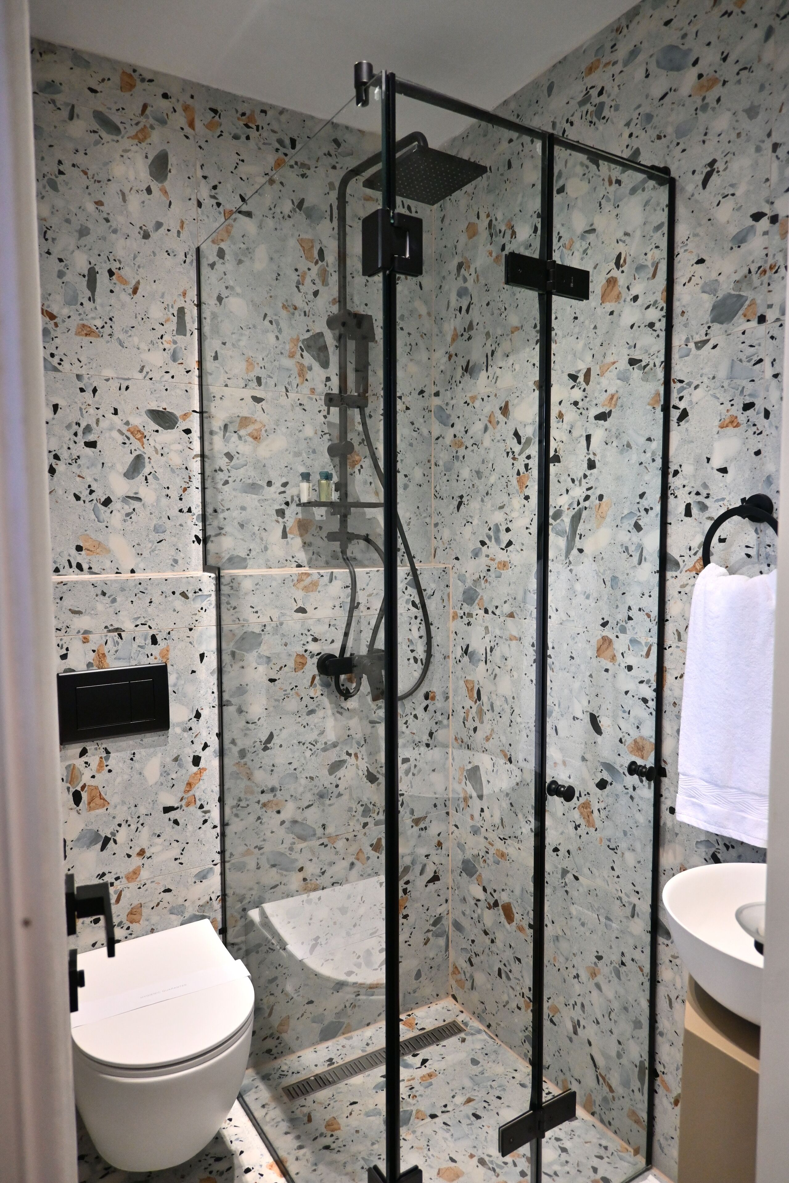 A modern bathroom with a glass-enclosed shower, a white toilet, and a sink with a towel hanging on the wall.