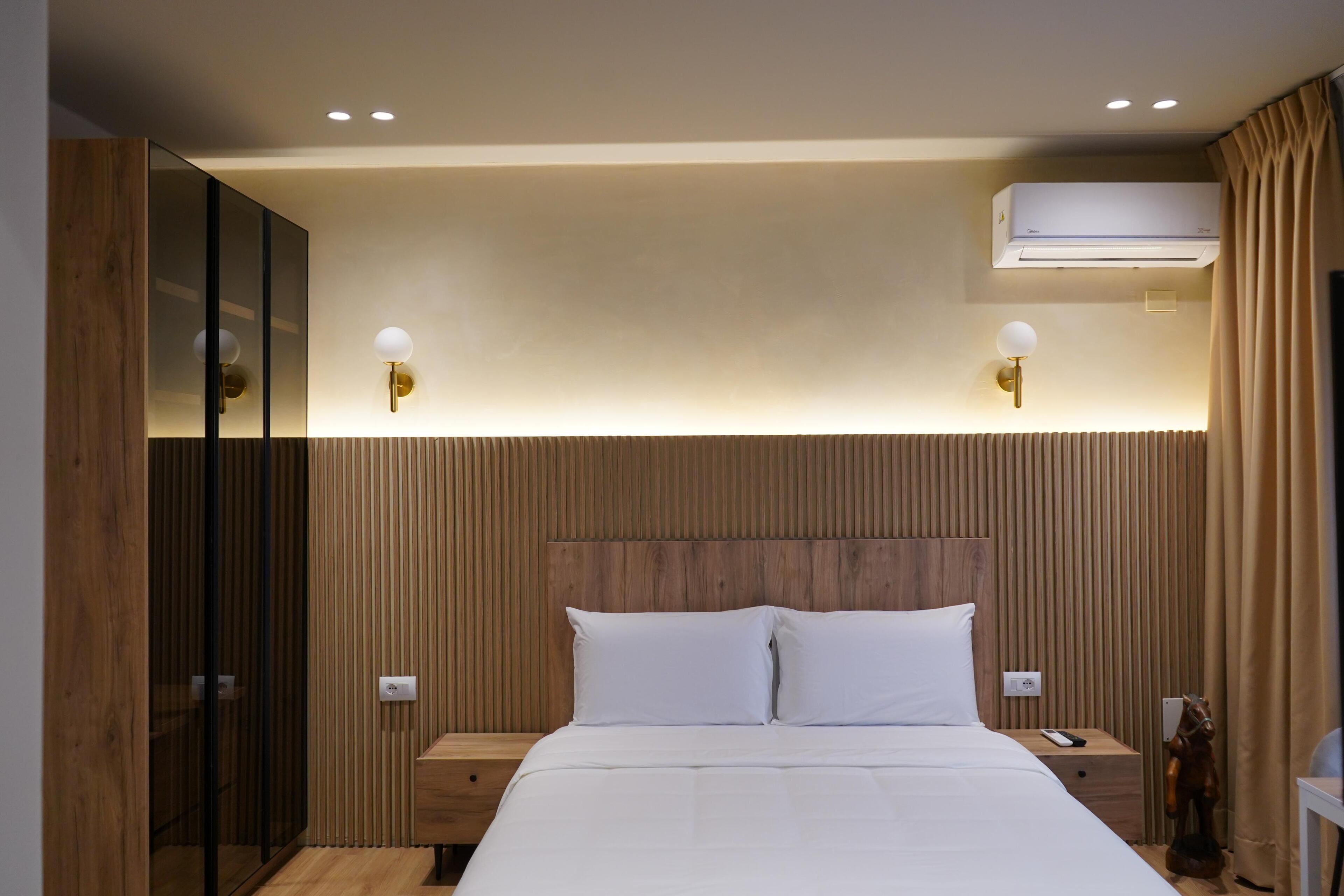 Een moderne hotelkamer met een houten hoofdbord, een groot bed met witte lakens, twee nachtkastjes met lampen en een airconditioner boven het bed.
