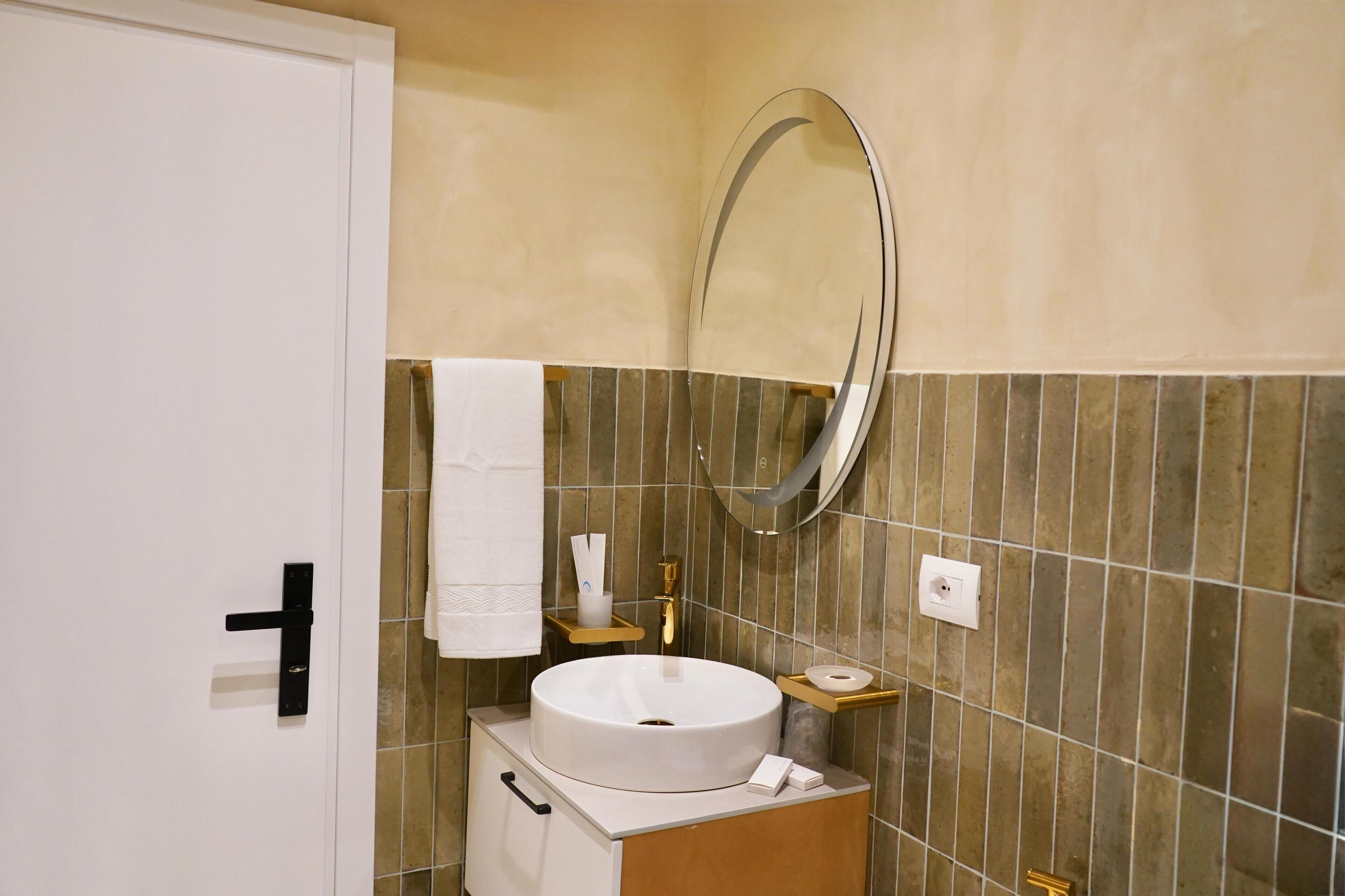 A bathroom with a white sink, a round mirror, a towel hanging on the wall, and a small shelf with toiletries.
