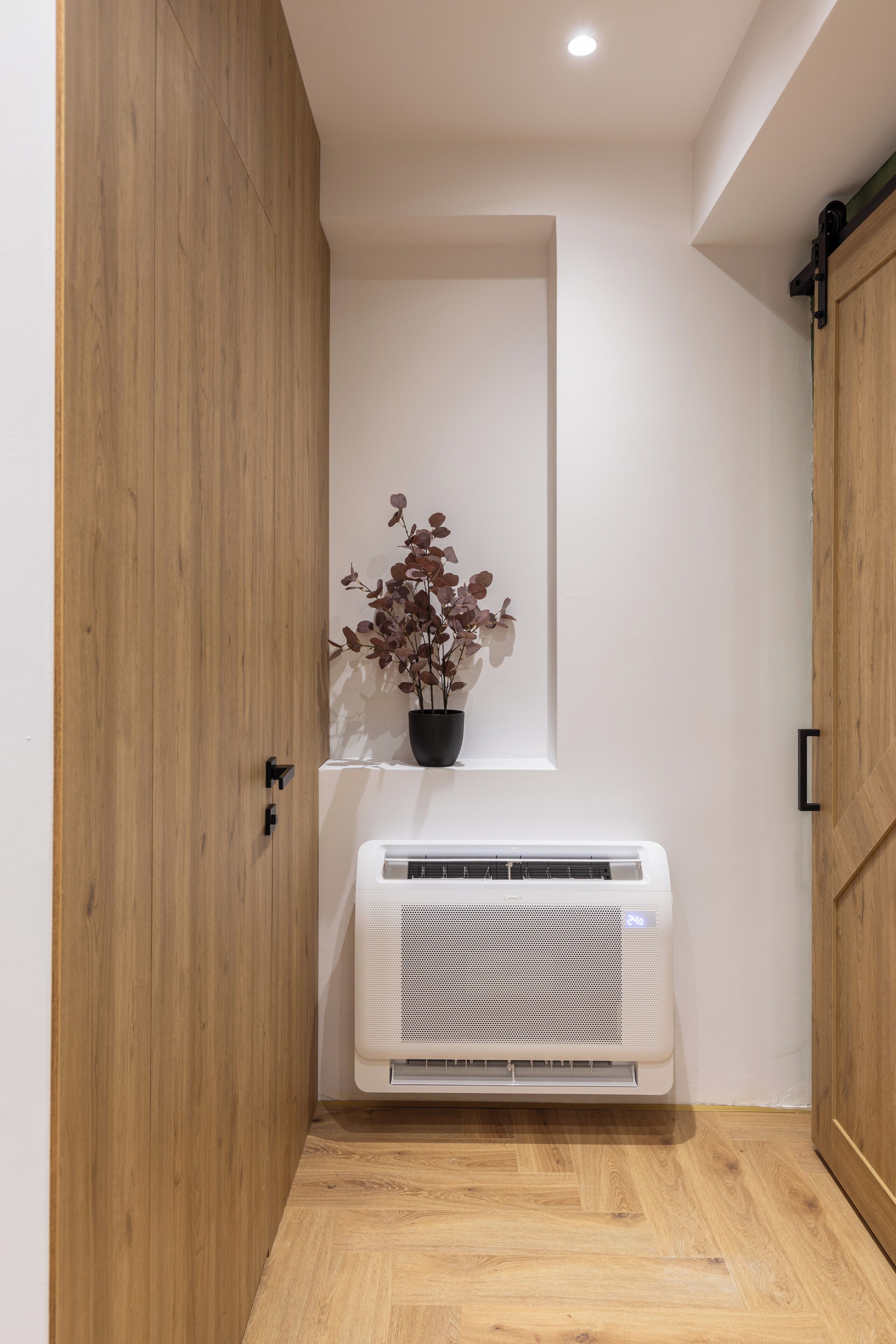 Una habitación con piso de madera, pared blanca, una planta en maceta y un equipo de aire acondicionado blanco.