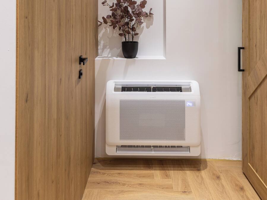 Una habitación con piso de madera, pared blanca, una planta en maceta y un equipo de aire acondicionado blanco.