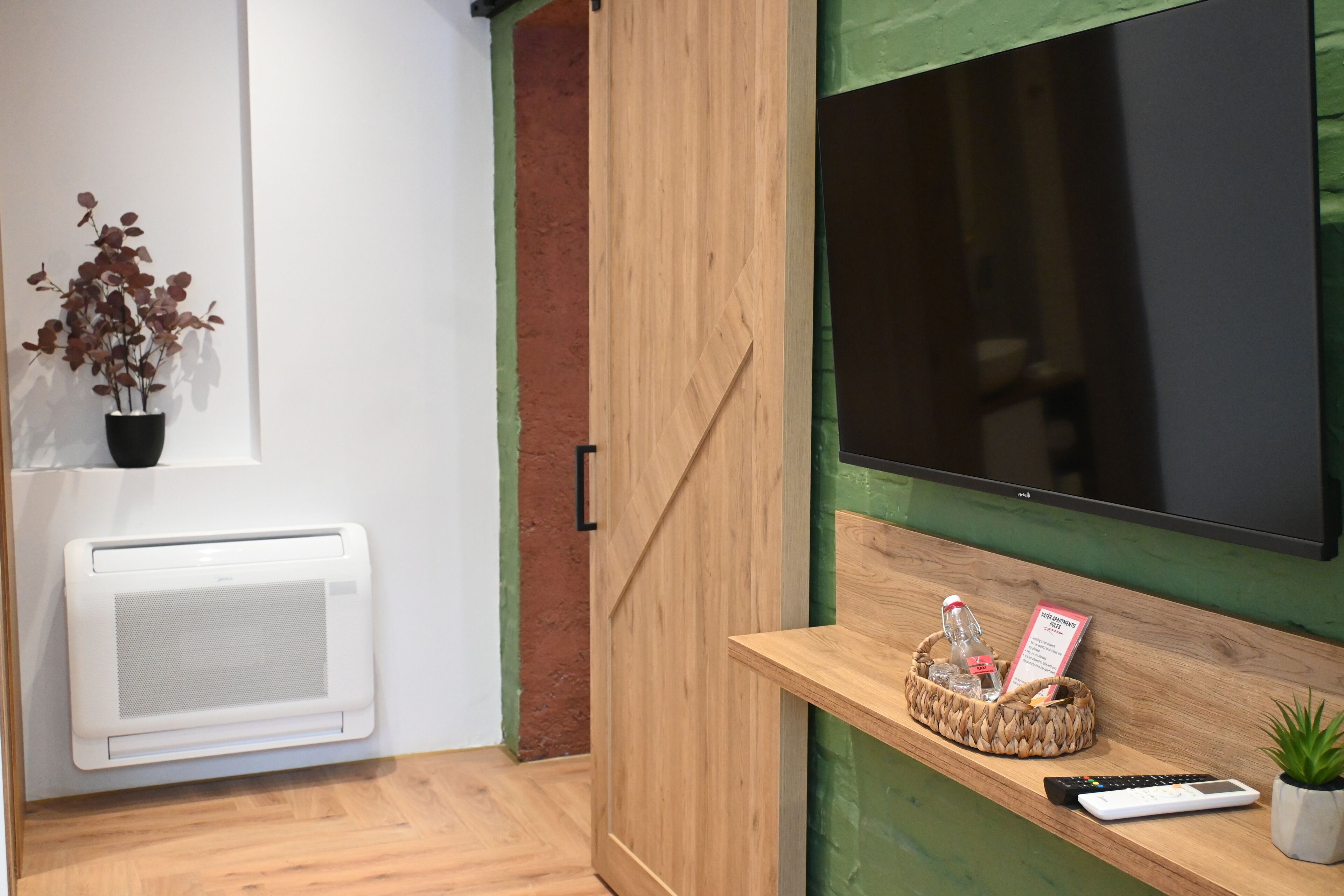 A room with a wooden cabinet, a mounted TV, a small shelf with a plant, a basket with a bottle, and a remote control.
