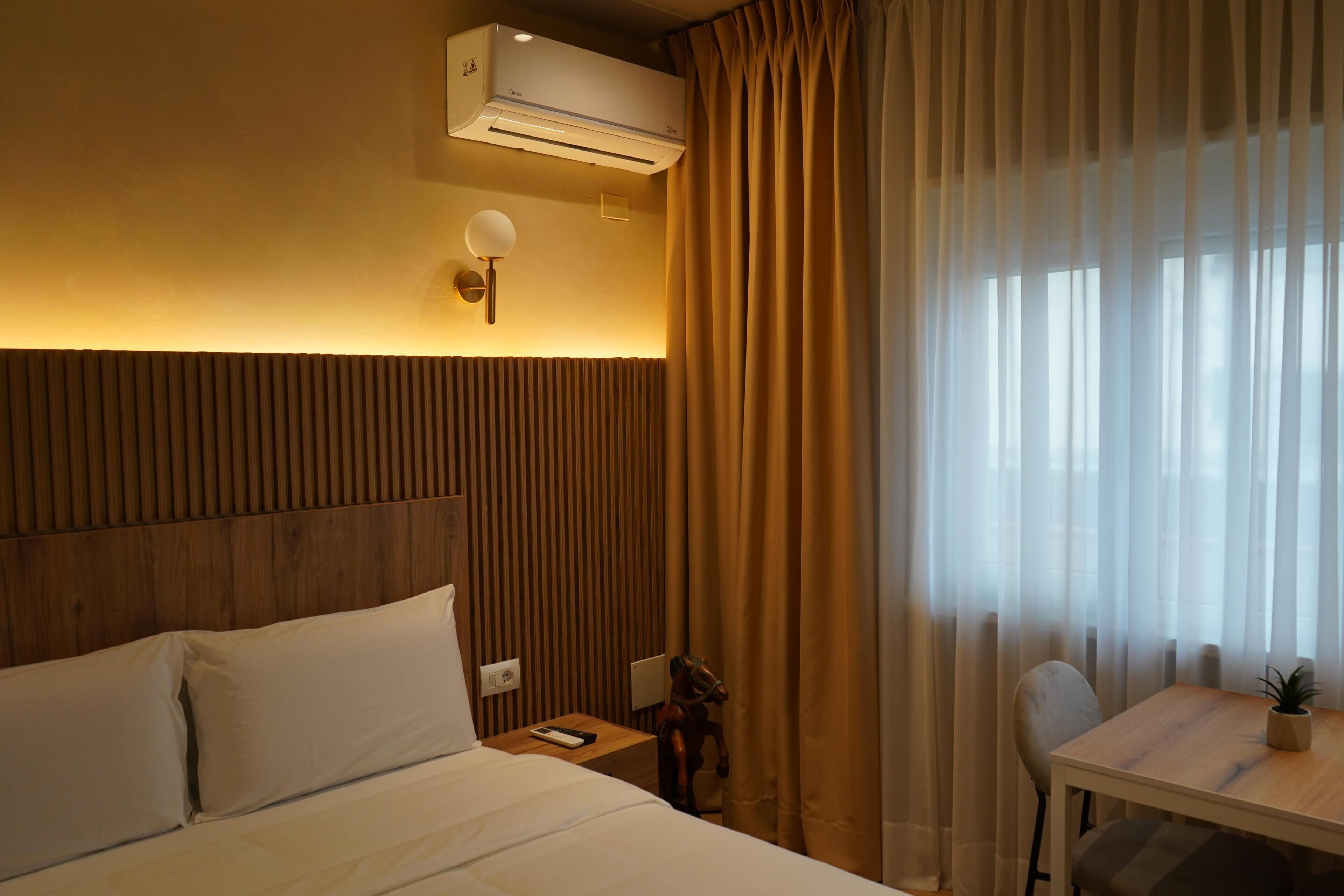 Hotelkamer met een houten hoofdbord, een bed met witte lakens, een kleine tafel met stoel, en een raam met dunne gordijnen.