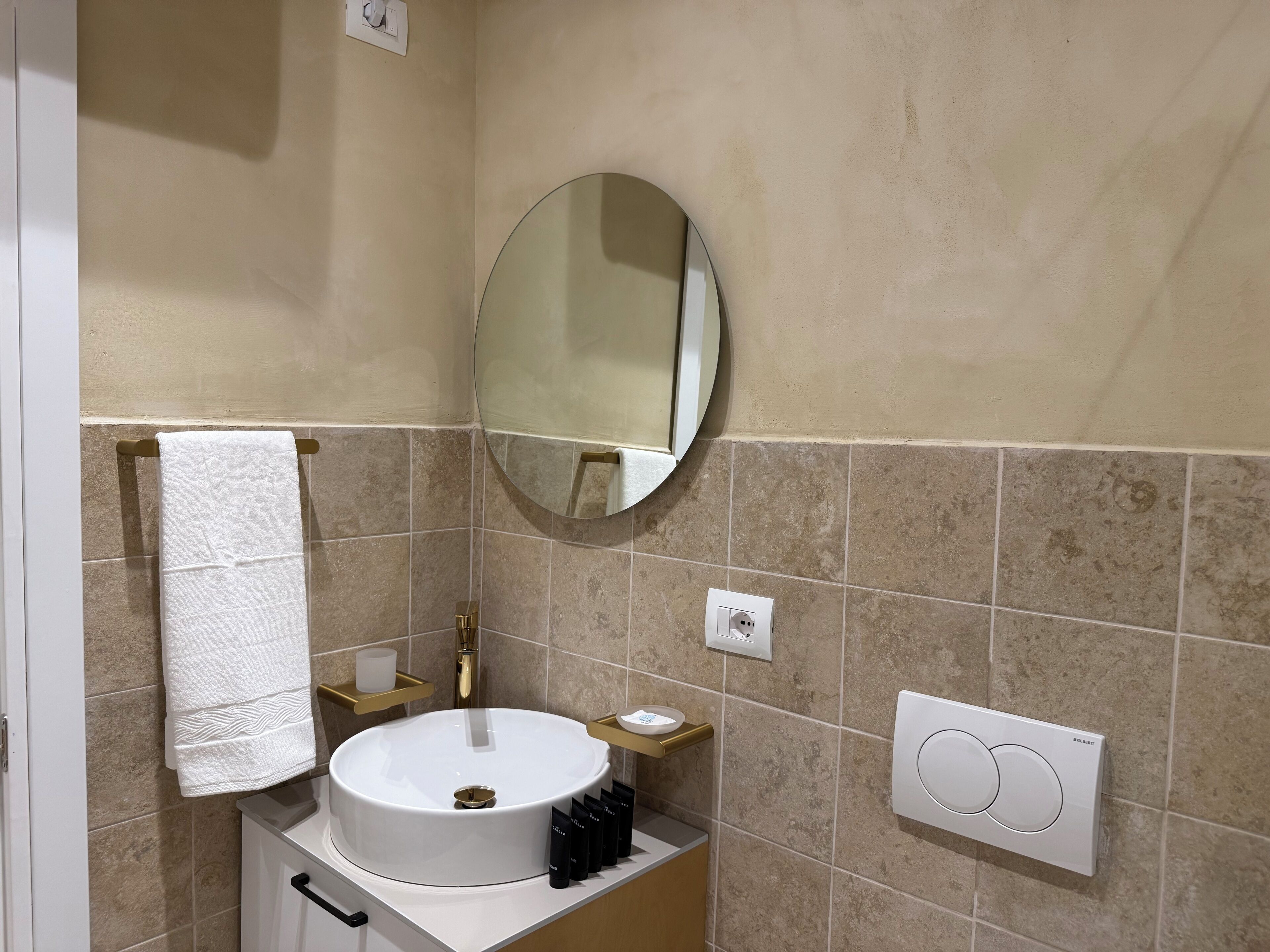 Un baño con un lavamanos blanco, un espejo redondo, un perchero para toallas, un dispensador de jabón e interruptor de luz.