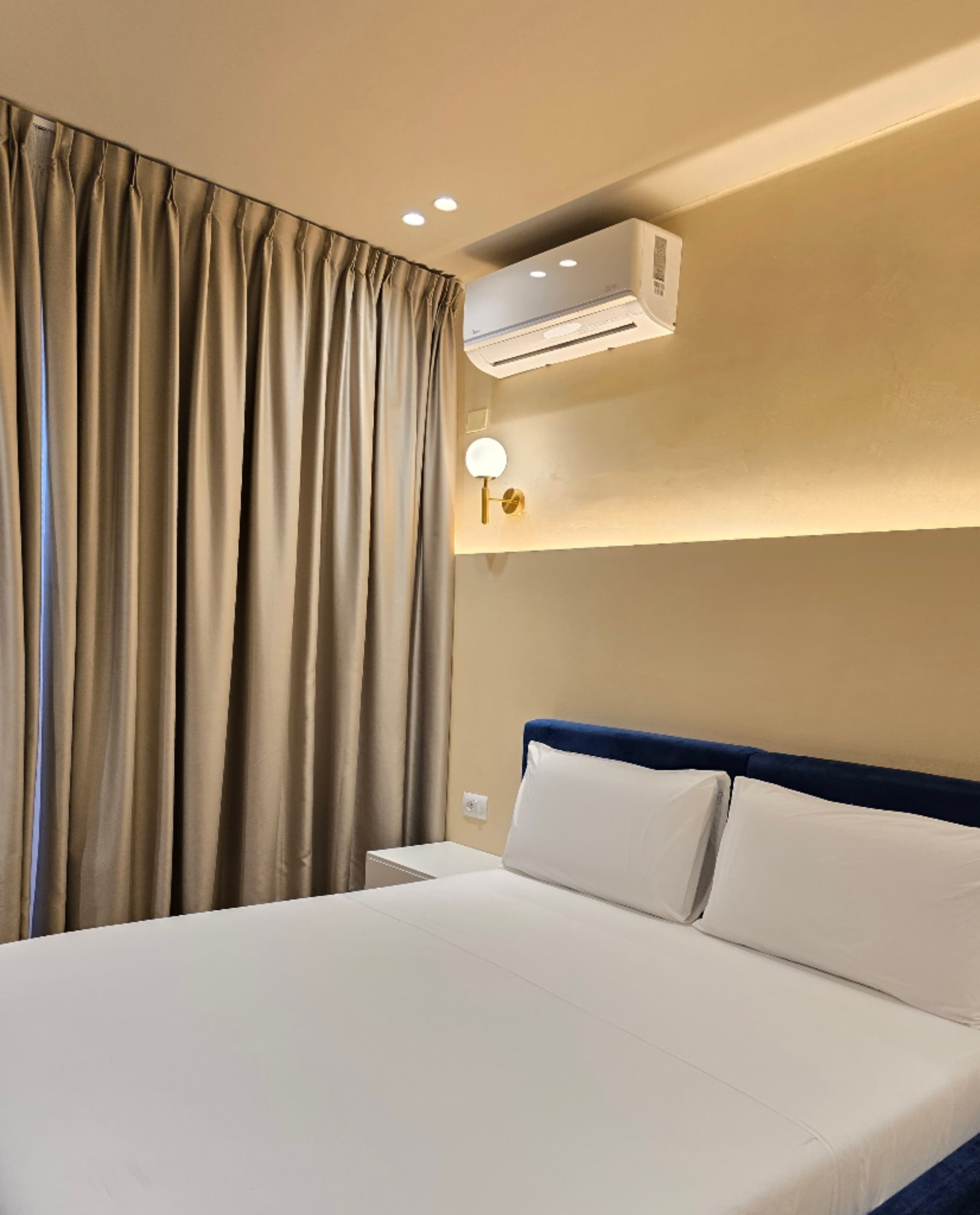 Une chambre d’hôtel avec un grand lit, des rideaux beiges, un climatiseur fixé au mur et une table de chevet avec une lampe.