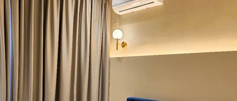 Una habitación de hotel con una cama grande, cortinas color beige, un aire acondicionado montado en la pared y una mesita de noche con lámpara.