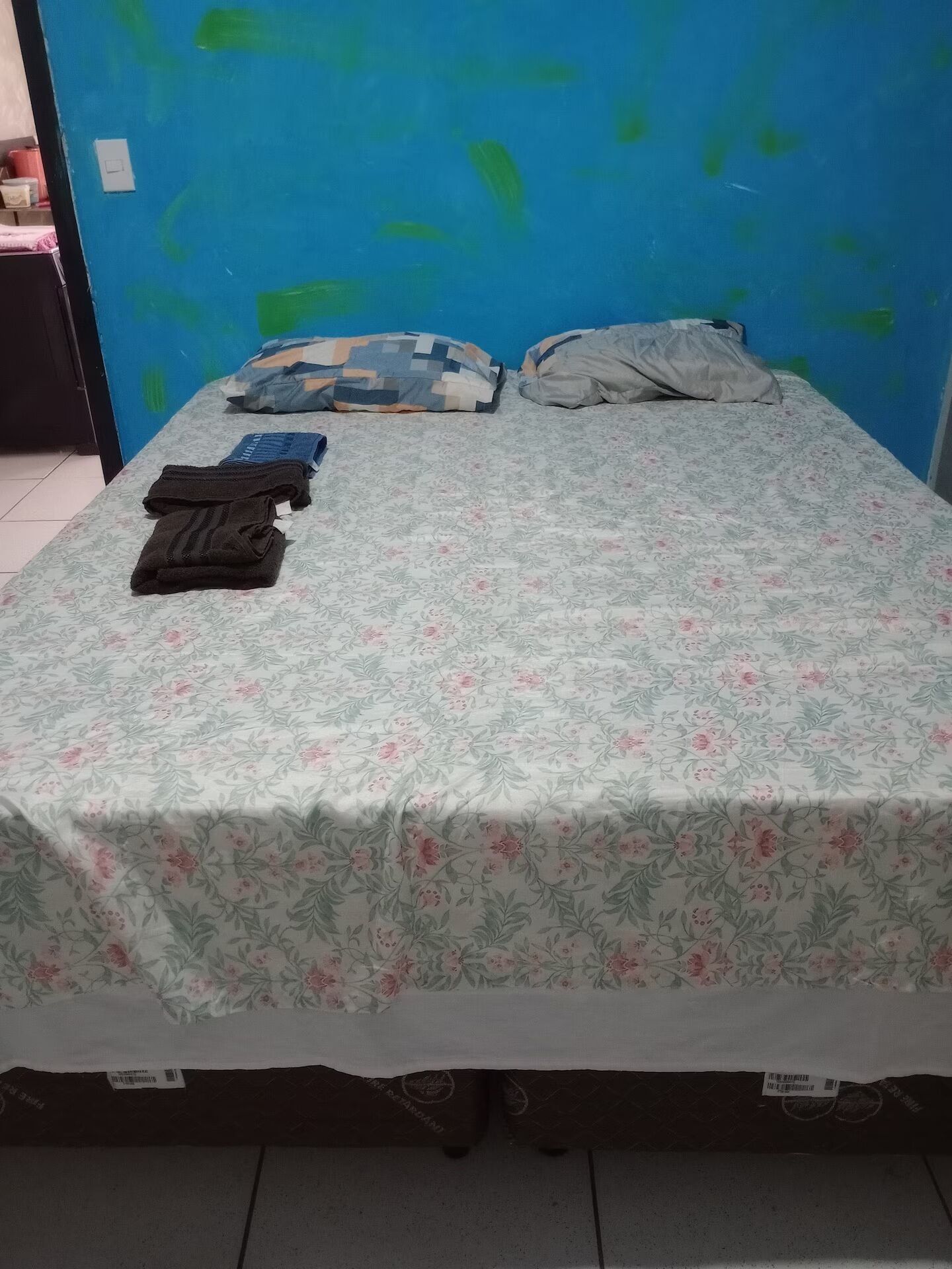 A bed with a floral bedspread, pillows, and folded clothes on a tiled floor.