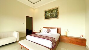 A hotel room with a bed, a bench, a wooden nightstand, a painting on the wall, and a door leading to another room. - Oemah Surya Ubud (Ubud)