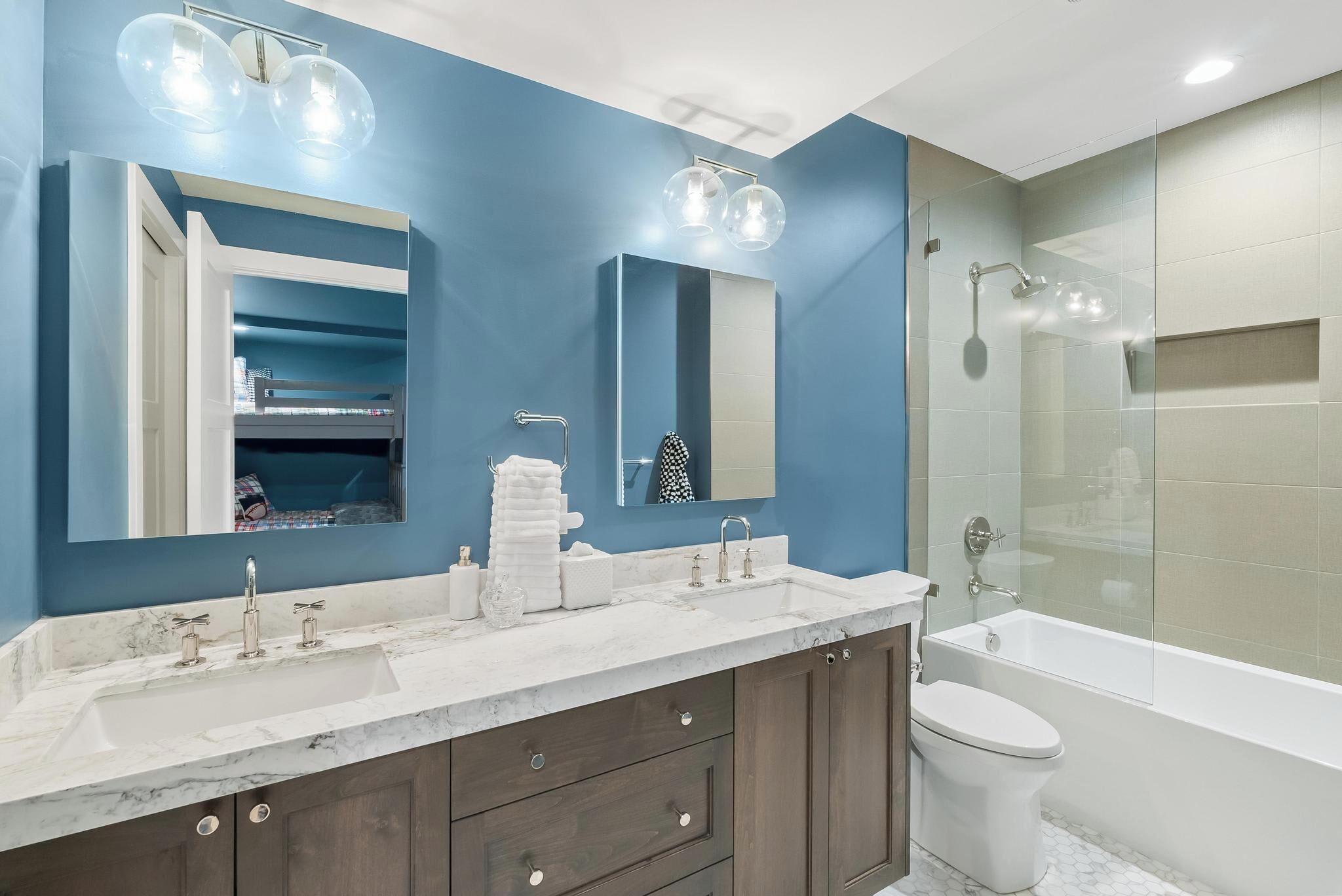 A modern bathroom with a blue accent wall, a large mirror, a double sink vanity, and a glass-enclosed shower area.