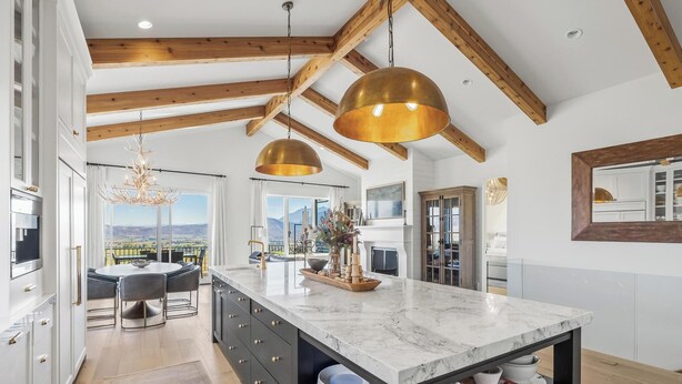 Cocina moderna con isla de mármol, vigas de madera, lámparas colgantes y vistas a las montañas.