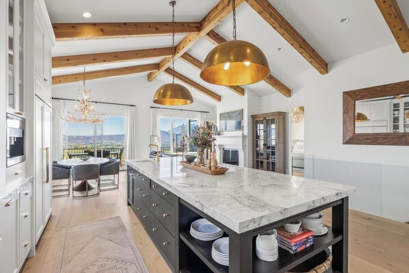 Cocina moderna con isla de mármol, vigas de madera, lámparas colgantes y vistas a las montañas.