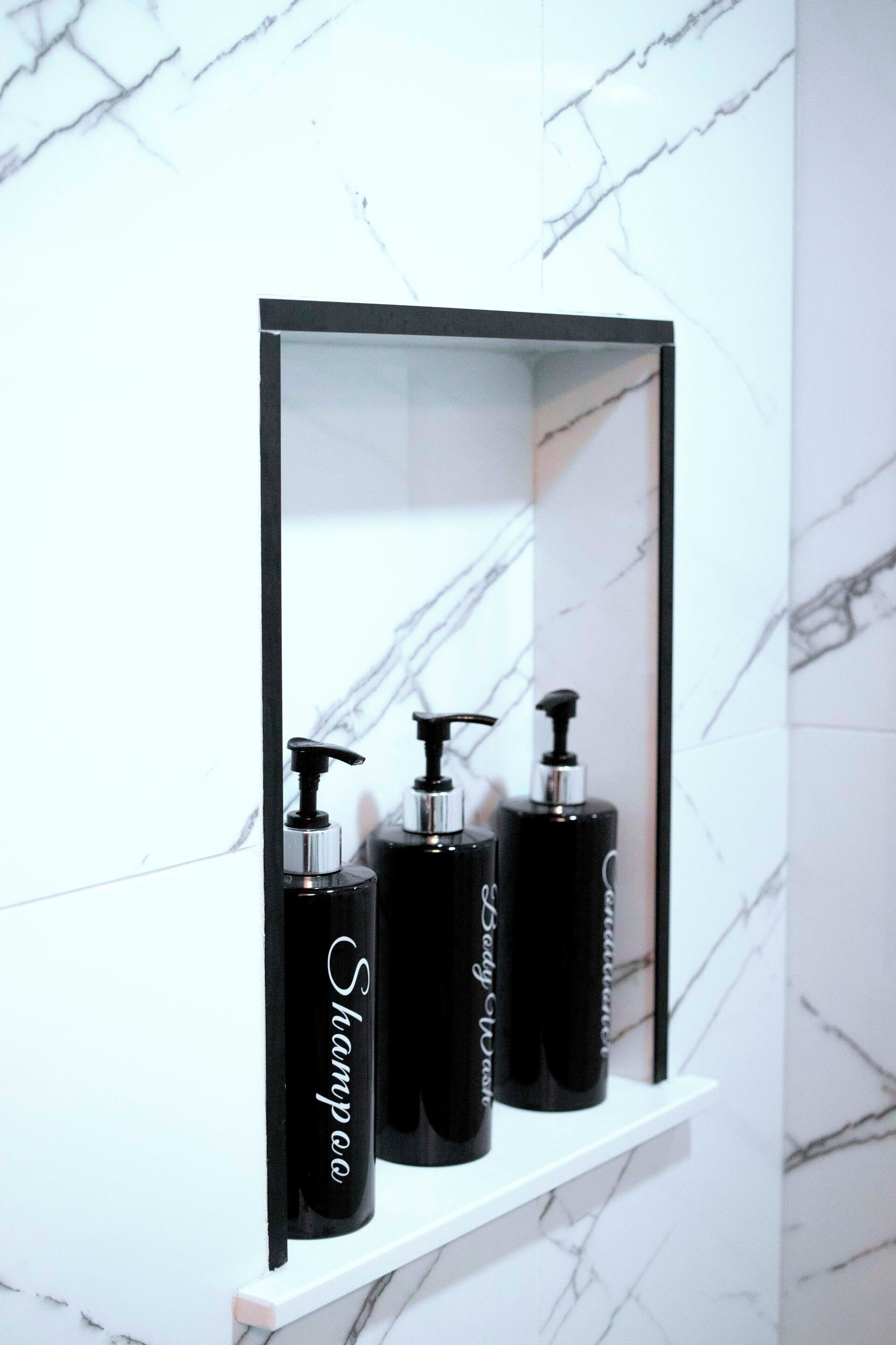 A bathroom with a white marble wall and a shelf holding three black bottles of shampoo.