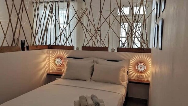 Een tweepersoonsbed met een netjes gevouwen handdoek, twee nachtkastjes met decoratieve lampen, en een geometrisch wandontwerp.