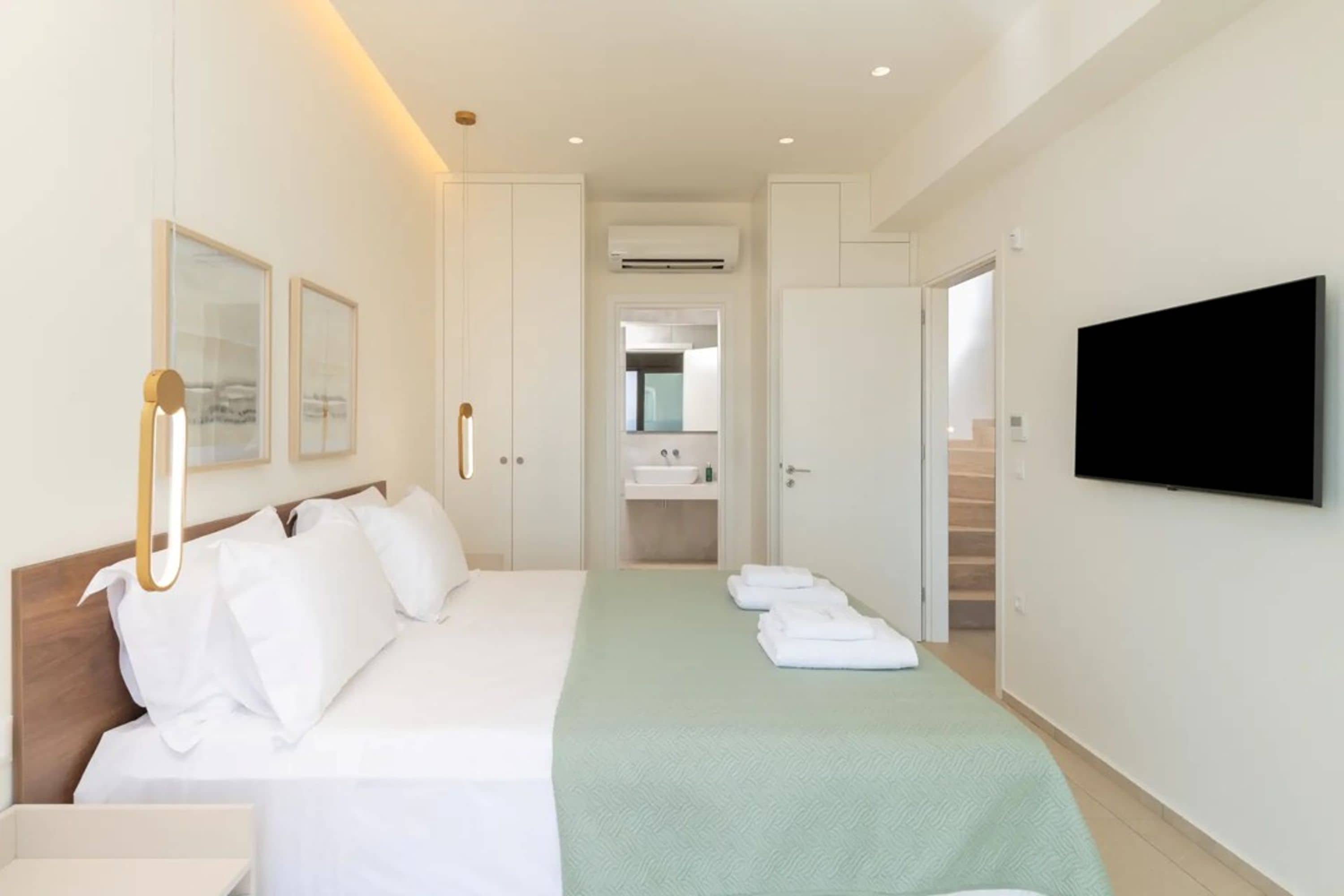 Habitación de hotel con cama, televisión, baño con lavamanos y espejo, y un pasillo que conduce a otra habitación.
