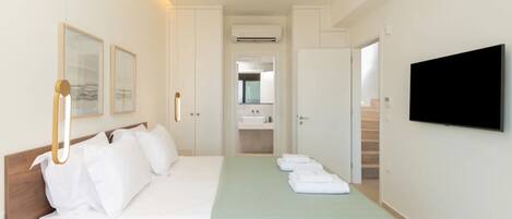 Habitación de hotel con cama, televisión, baño con lavamanos y espejo, y un pasillo que conduce a otra habitación.