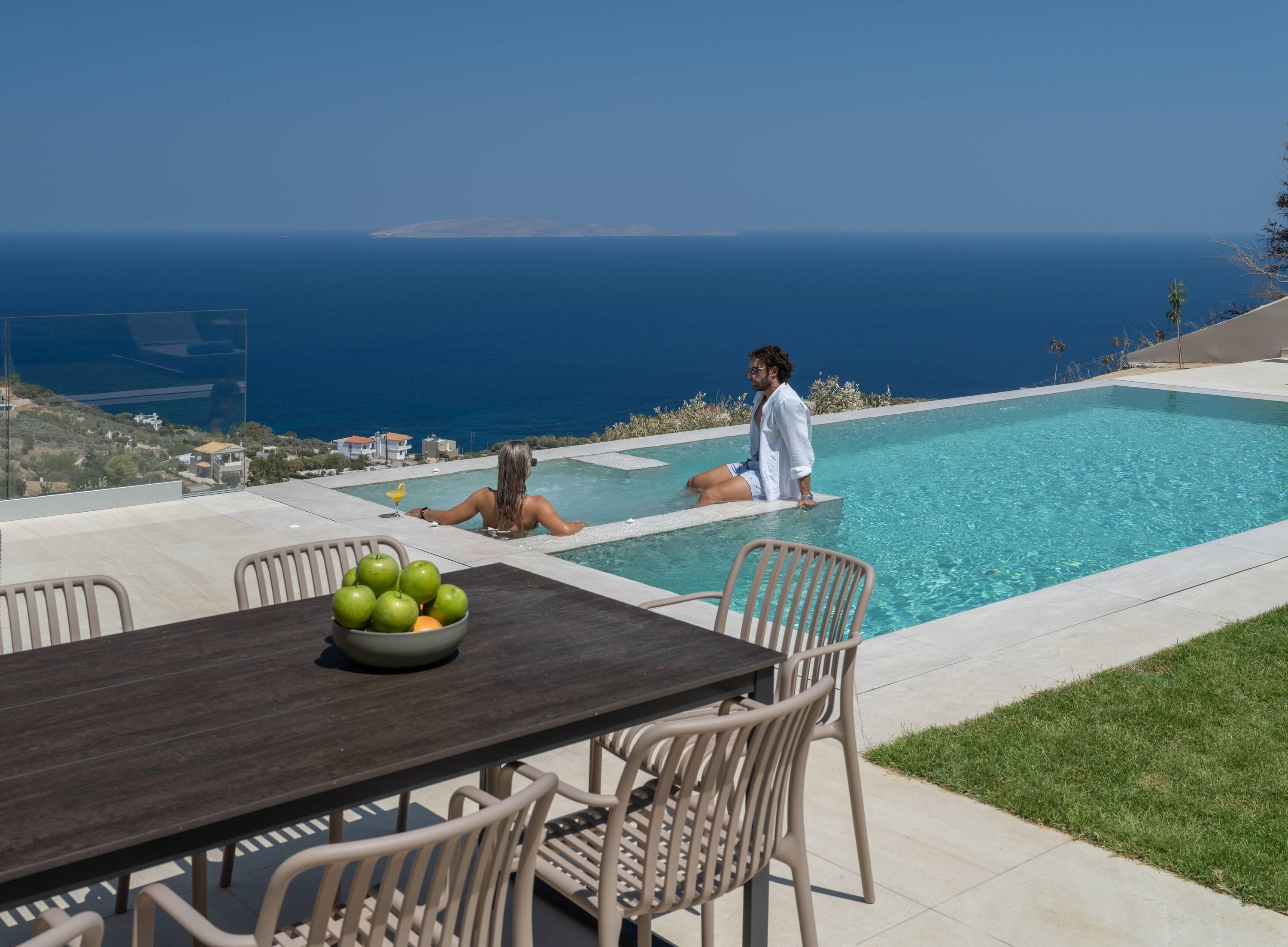 A poolside area with a table and chairs, a bowl of fruit, and a view of the sea.
