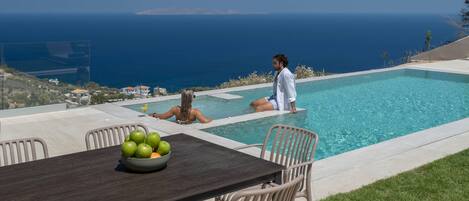 A poolside area with a table and chairs, a bowl of fruit, and a view of the sea.
