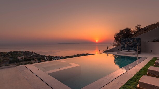 A modern outdoor pool area with a clear view of the sunset over the sea, a stone wall, and lounge chairs. - Luxury Villa Vie (Malevizi)