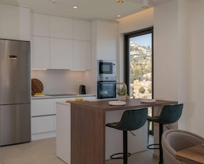 A modern kitchen with white cabinetry, a stainless steel refrigerator, a built-in oven, and a microwave. There is a wooden island with two bar stools and a view of a mountainous landscape through a large window.