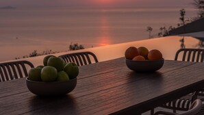A wooden table with two bowls of fruit, one with green apples and the other with oranges, set against a sunset view over a calm body of water. - Luxury Villa Vie (Malevizi)