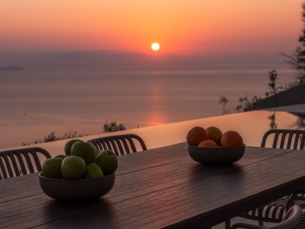 A wooden table with two bowls of fruit, one with green apples and the other with oranges, set against a sunset view over a calm body of water.