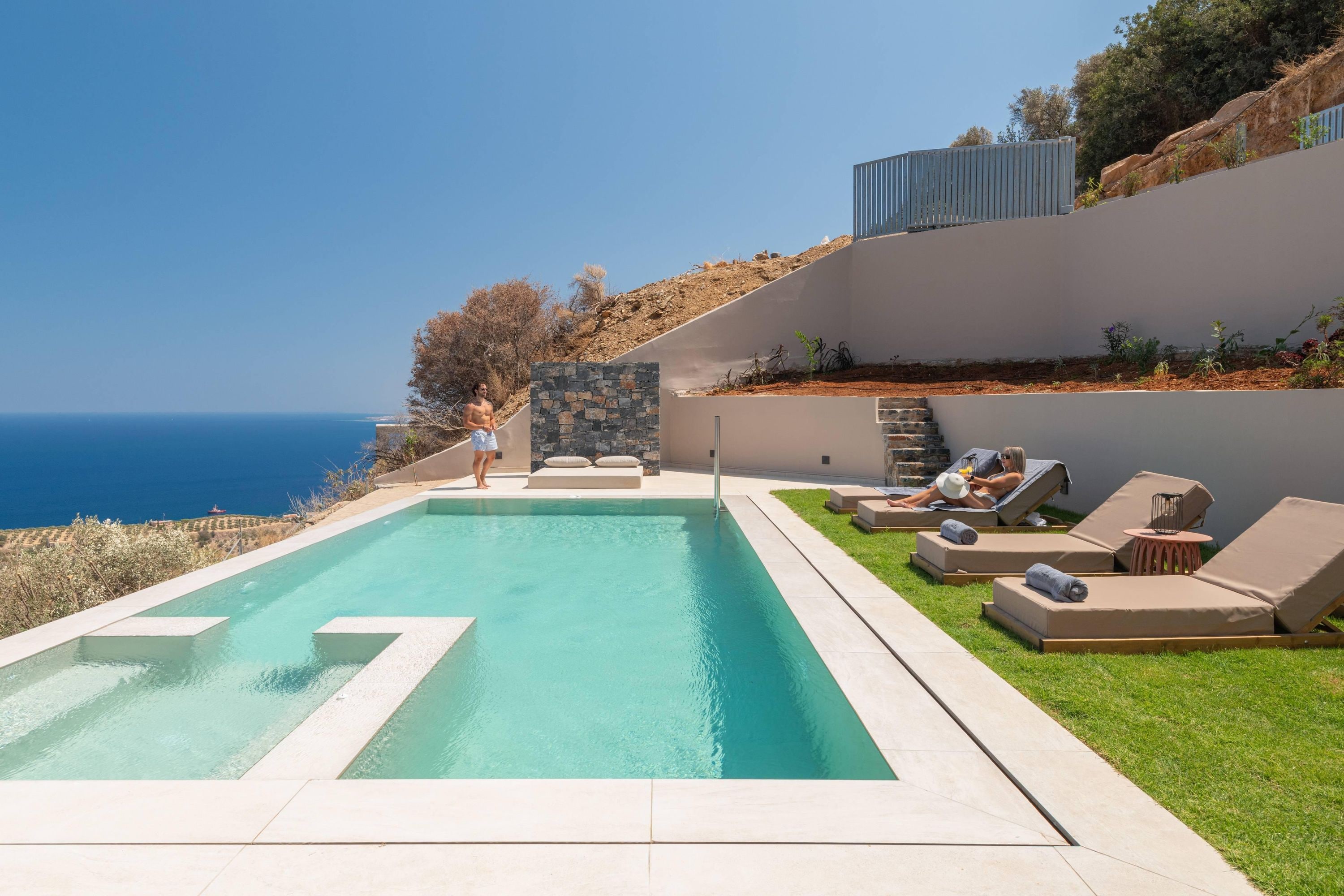 Une piscine offrant une vue dégagée sur l’océan, entourée de chaises longues et d’un mur en pierre.