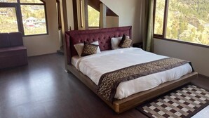 A large bed with a wooden frame and a headboard, positioned in a room with a view of a mountainous landscape through large windows. - Hotel Apple View Narkanda (Theog)