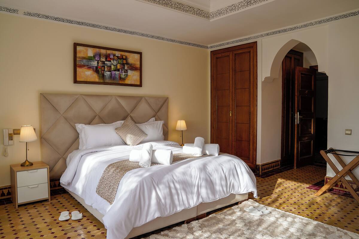 A neatly made bed with white linens and a decorative headboard, a bedside lamp, and a pair of slippers on the floor.