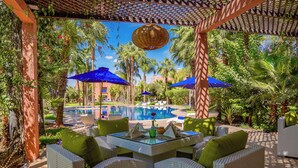 An outdoor seating area with wicker furniture and blue umbrellas, overlooking a pool and lush greenery. - Palais Volteras (Marrakech)