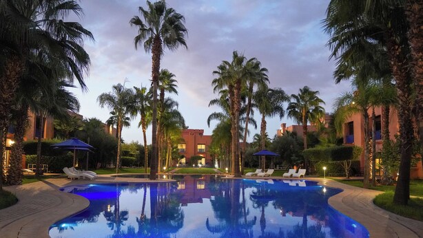 A swimming pool surrounded by palm trees and lounge chairs, with buildings in the background. - Palais Volteras (Marrakech)