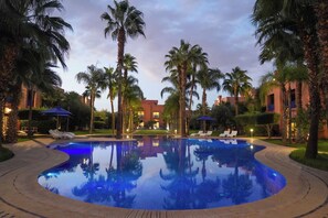 A swimming pool surrounded by palm trees and lounge chairs, with buildings in the background. - Palais Volteras (Marrakech)