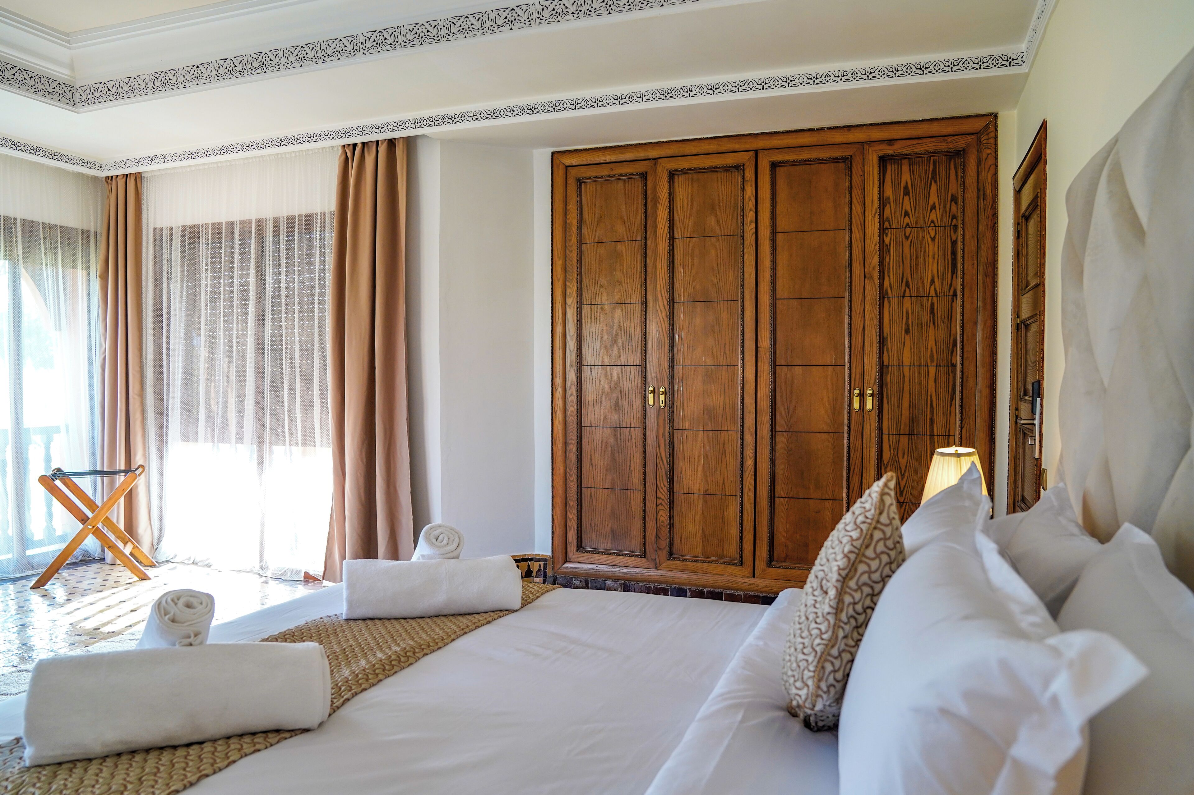 A hotel room with a large bed, a wooden wardrobe, a sitting area with a chair and a table, and a view of the outdoors.