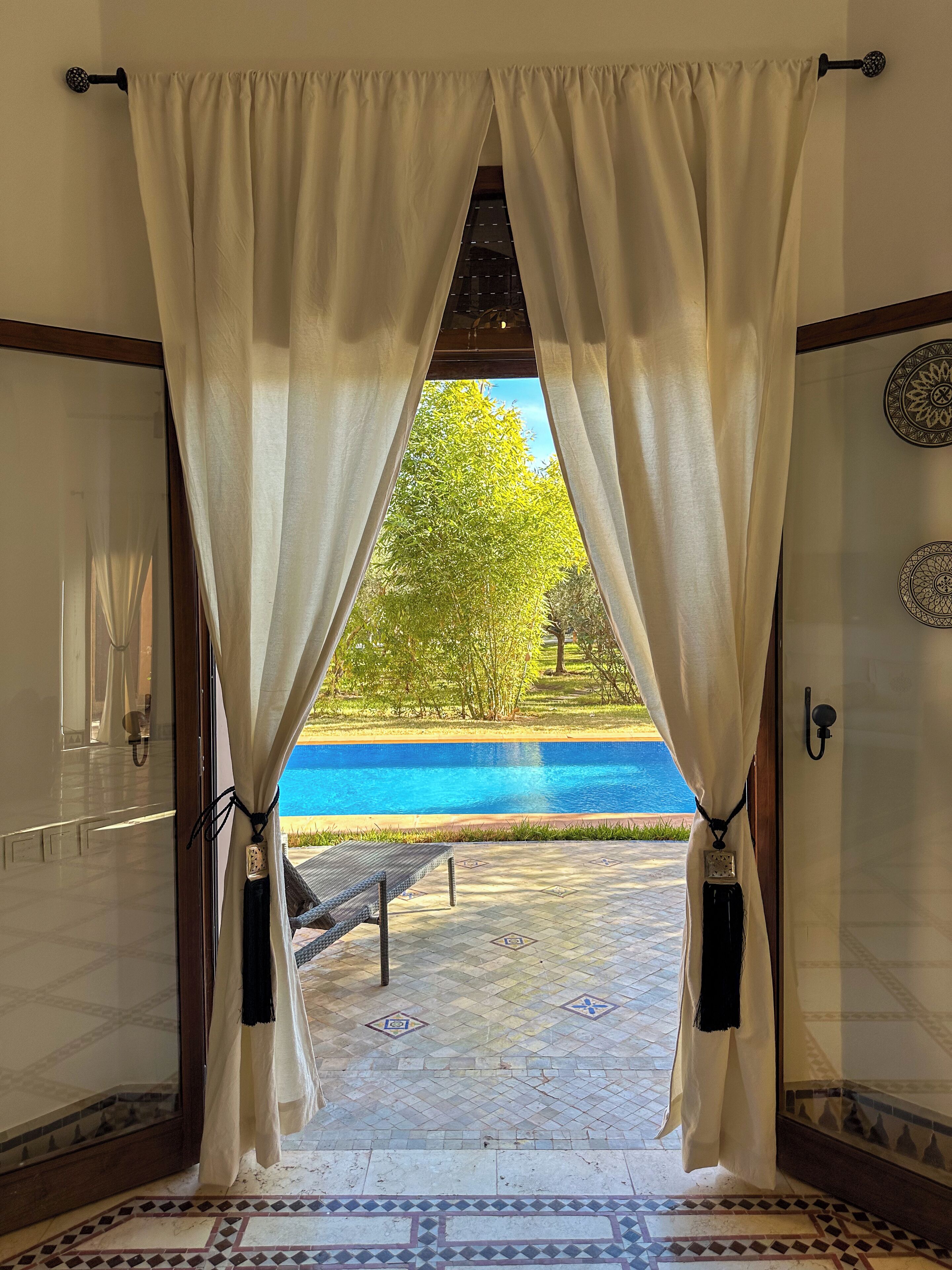 A view through a doorway with white curtains, leading to a pool area with a tiled floor and a bench.