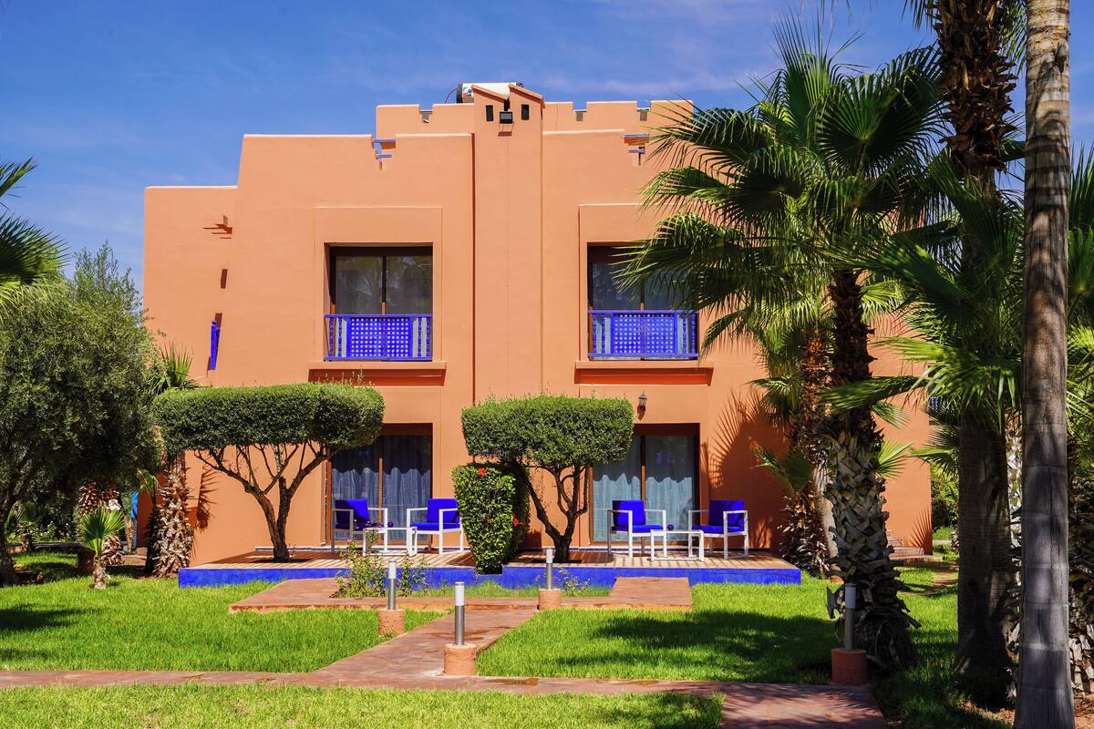 A building with a flat roof, two balconies with blue railings, and a well-maintained lawn with palm trees.