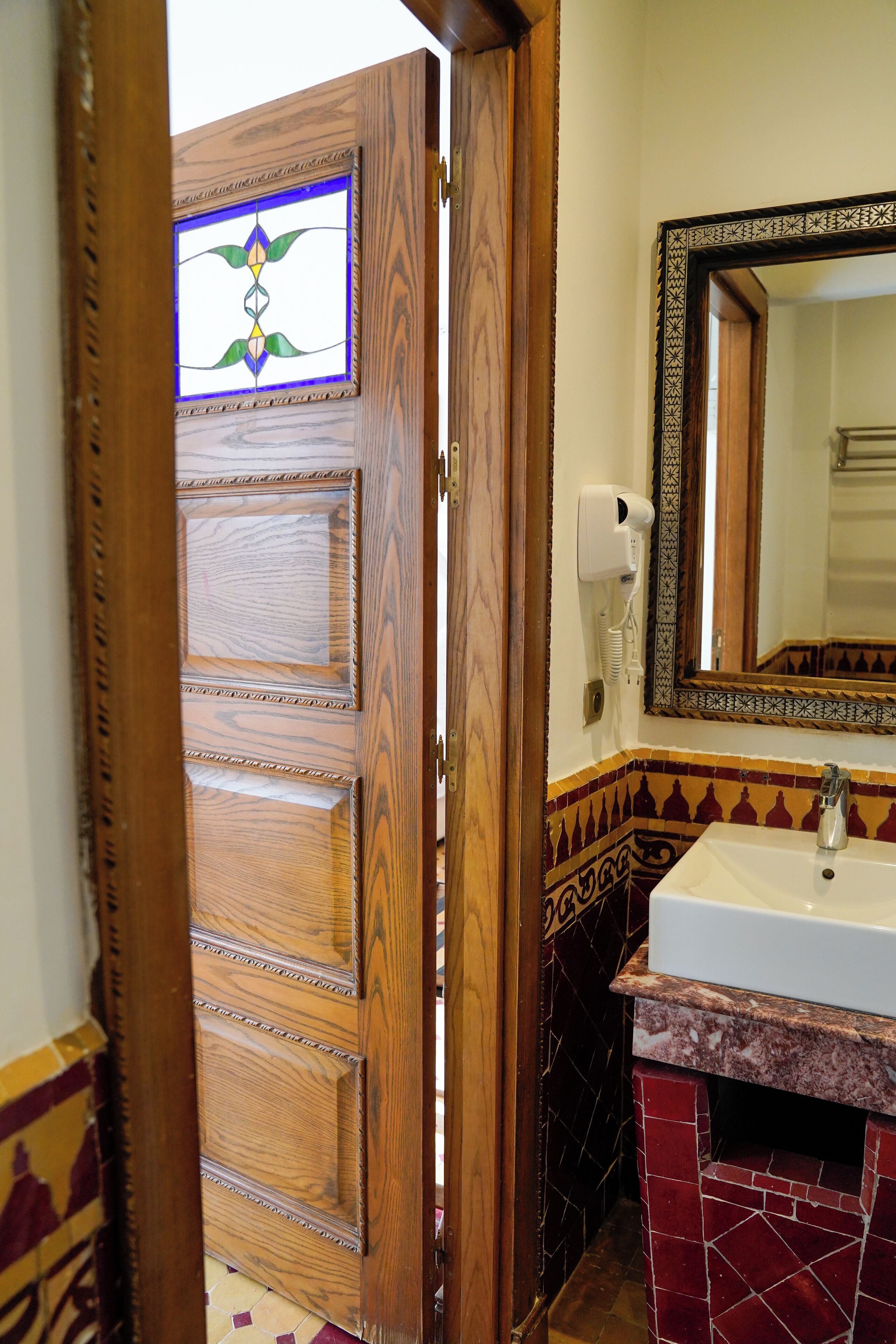 Una puerta de madera con un vitral, un baño con un lavamanos blanco y un espejo, y un suelo de baldosas.
