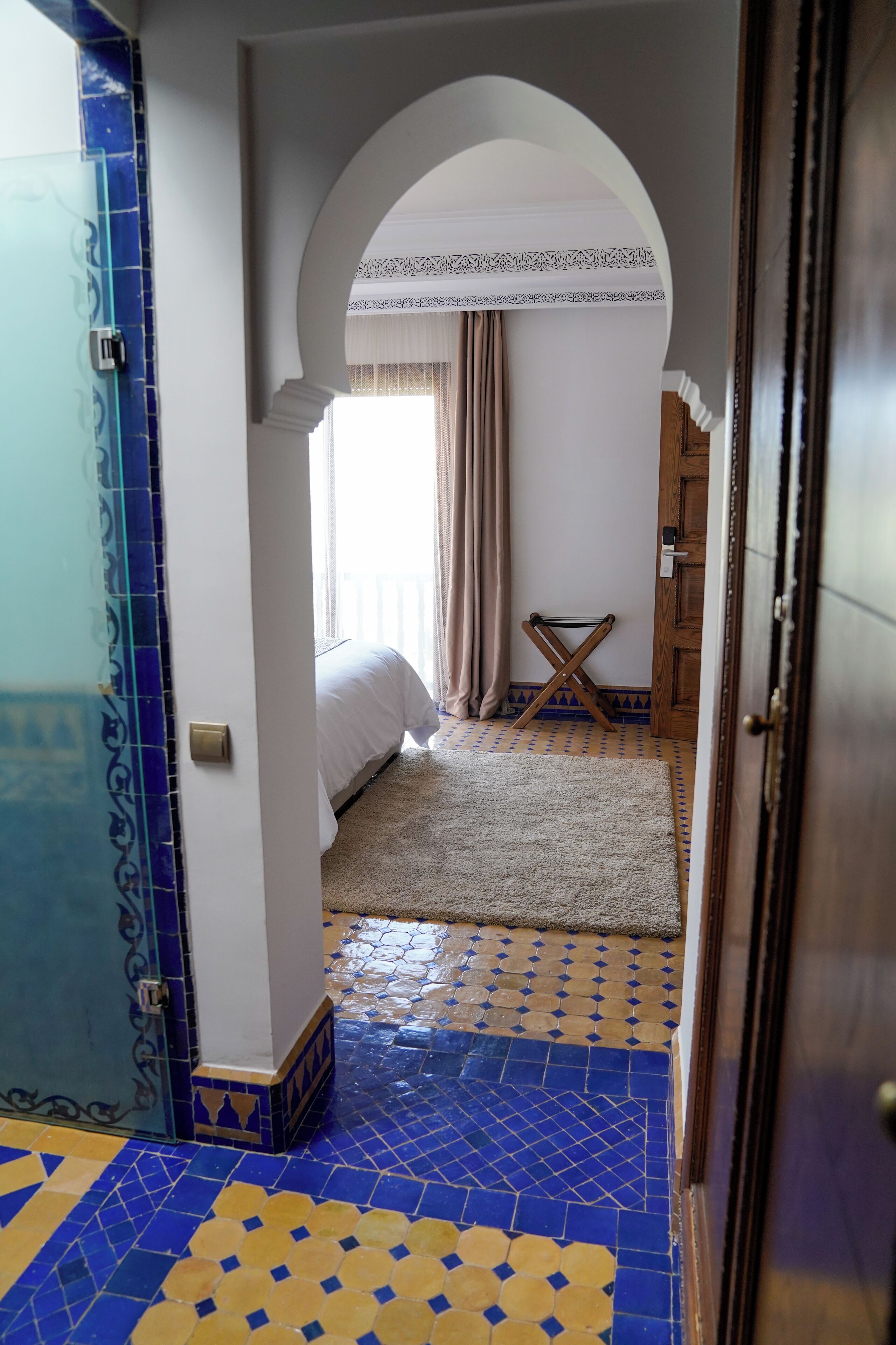 A room with a patterned blue and yellow floor, a bed with white bedding, a small table, and a window with curtains.