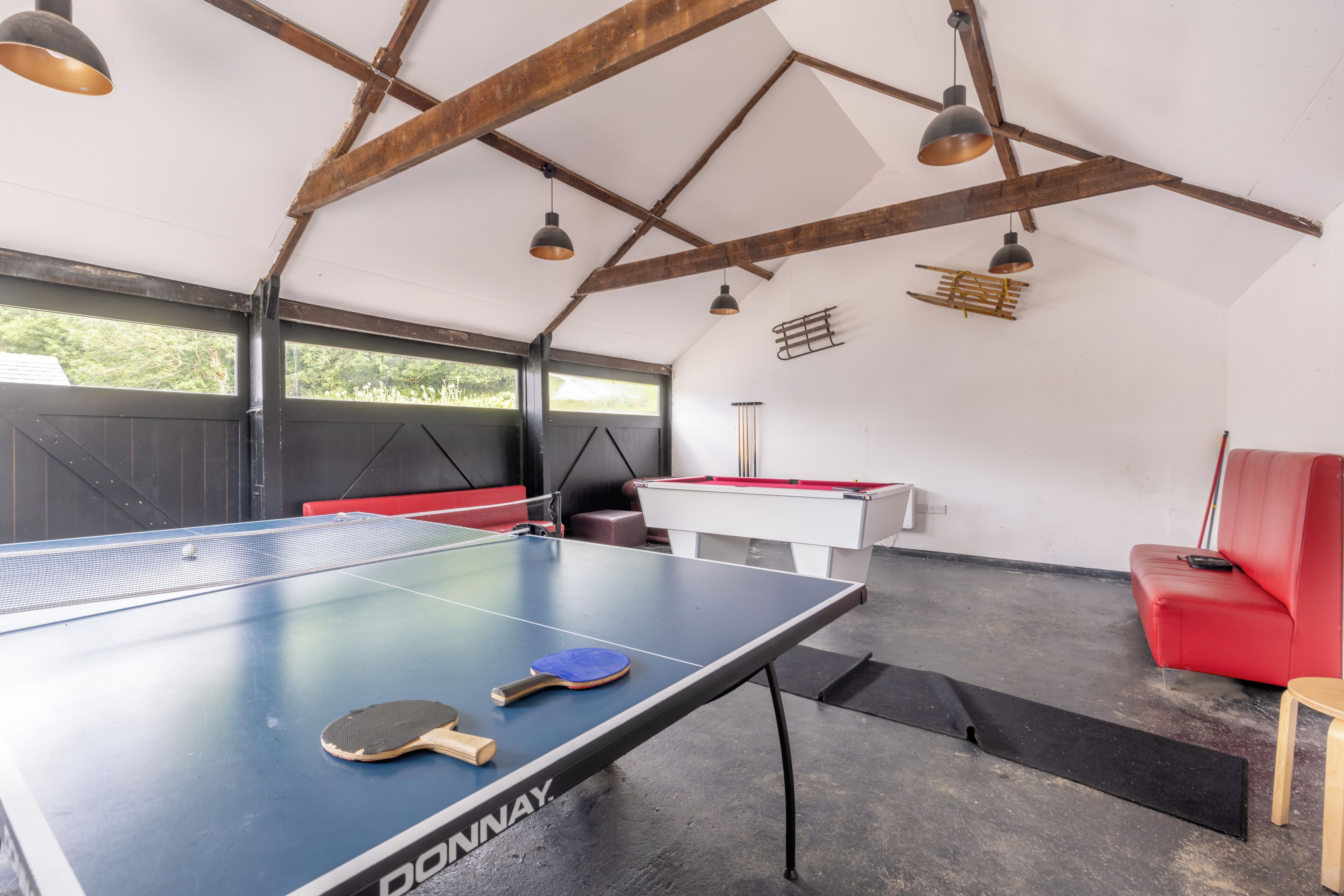 A ping pong table with two paddles, a red sofa, and a pool table in a room with exposed wooden beams and large windows.
