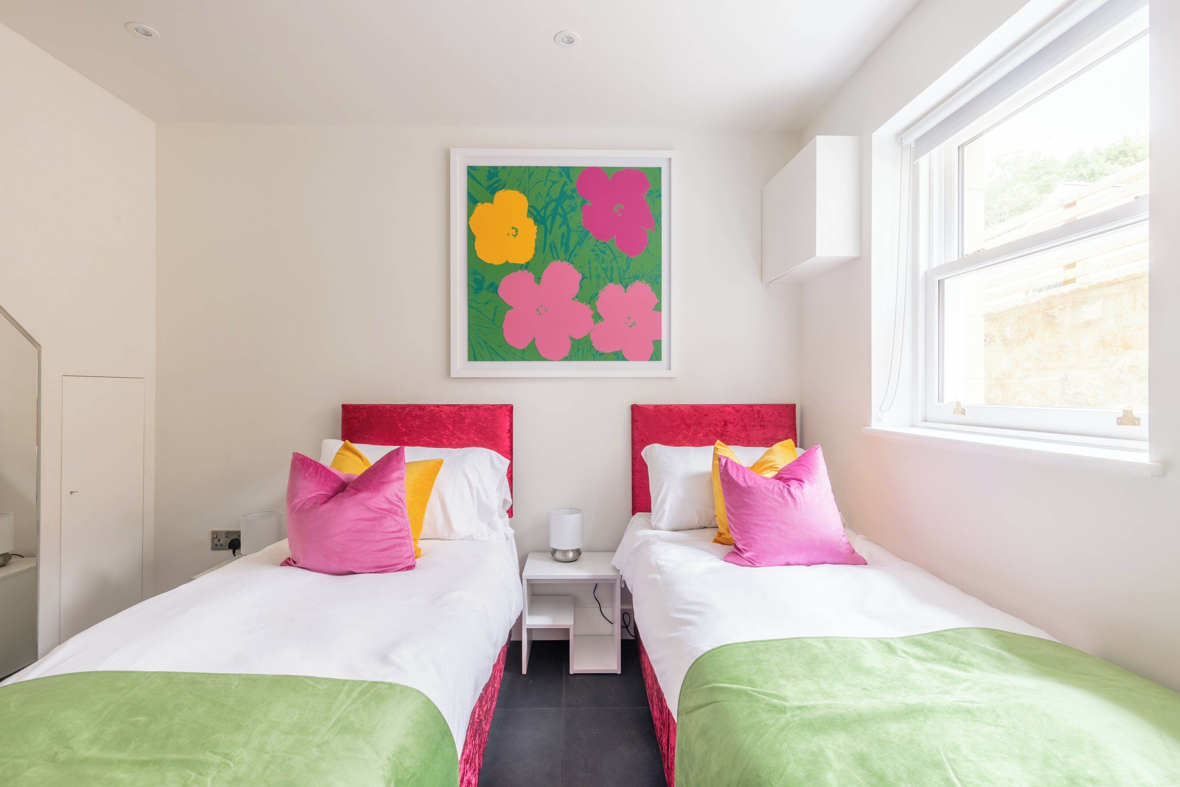 A hotel room with two beds, each with a red headboard, white bedding, and colorful pillows. There is a small bedside table with a lamp, a green wall art piece above the beds, and a window on the right side.