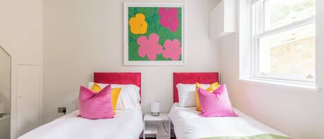 A hotel room with two beds, each with a red headboard, white bedding, and colorful pillows. There is a small bedside table with a lamp, a green wall art piece above the beds, and a window on the right side.