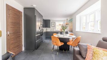 Una cocina moderna con armarios grises, una zona de comedor con una mesa redonda y sillas naranjas, y un sofá marrón con un cojín colorido.