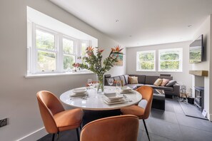 A modern dining area with a round table set for four, surrounded by orange chairs. There are large windows providing natural light and a view of greenery outside. A sofa and a fireplace are visible in the background. - Babbling Burn  - North East Escapes (Belford)