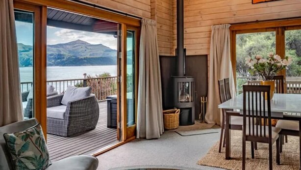 A wooden cabin with a fireplace, a dining area, and a balcony with a view of mountains and water.