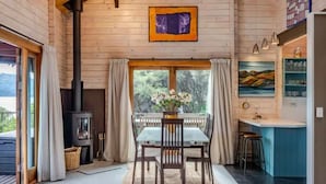 A wooden cabin interior with a dining table, chairs, a glass-top table, a fireplace, a kitchen area, and a large window.