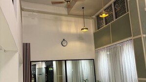 Een ruime woonkamer met hoge plafonds, een eettafel met stoelen, een groot raam met dunne gordijnen en een decoratieve klok aan de muur.