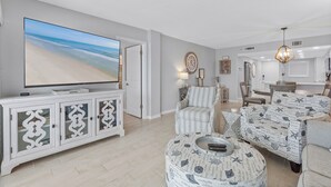 A modern living room with a large flat-screen TV, a striped armchair, a patterned ottoman, and a dining area in the background.