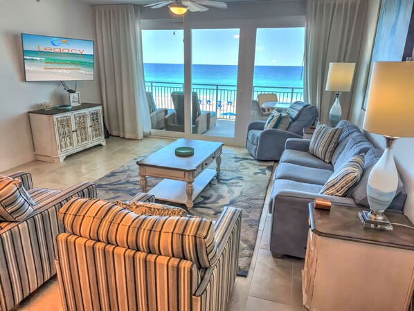 A spacious living room with a view of the ocean, featuring a sofa, chairs, a coffee table, and a television displaying 'Legacy Vacations'.